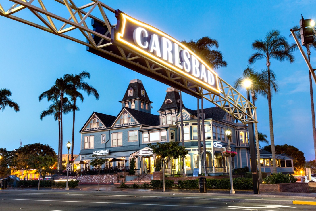 Illuminated Carlsbad gateway sign spanning a street beside palm trees and a Victorian-style building at dusk.