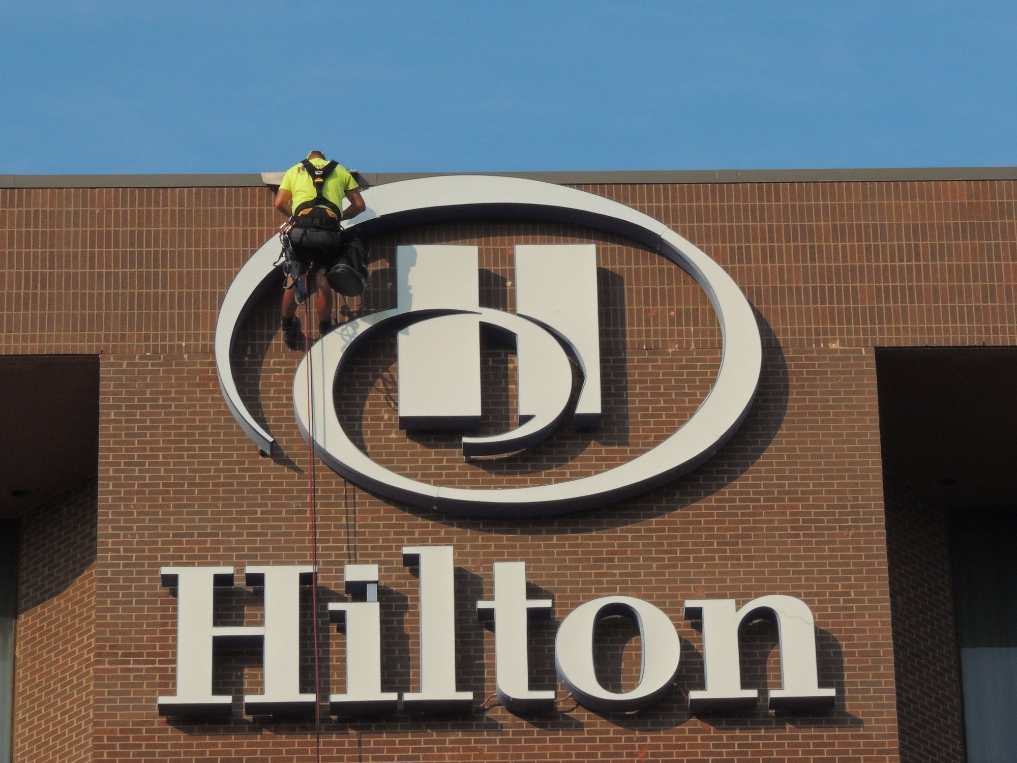 Sign technician suspended by ropes working on a large Hilton logo above dimensional lettering on a brick hotel exterior.