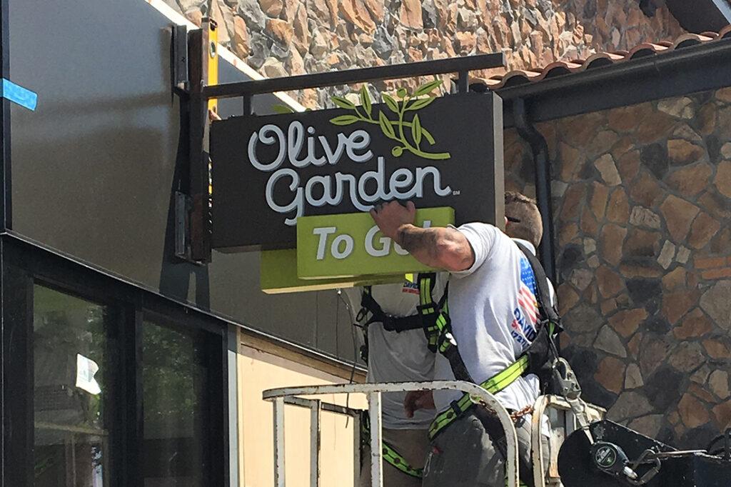 Sign technician in a bucket lift working on an Olive Garden To Go sign mounted beside the restaurant’s stone exterior.