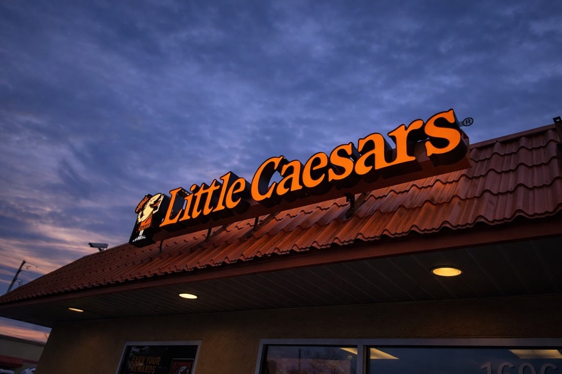 Illuminated orange Little Caesars letters and mascot logo above a tiled restaurant roof at dusk.