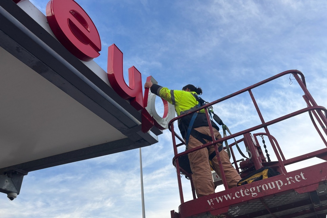 Sign technician wearing a safety harness in a boom lift working on red channel letters mounted above a storefront.