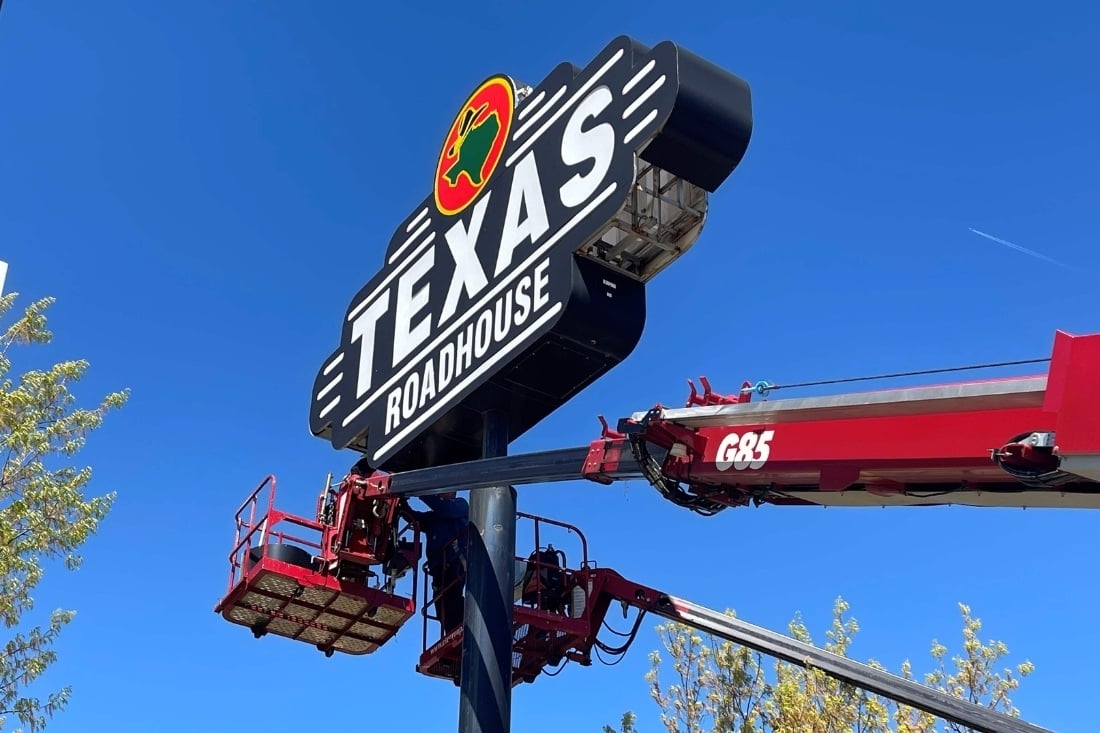 Technicians in aerial lifts servicing a Texas Roadhouse pole sign beneath a clear blue sky.