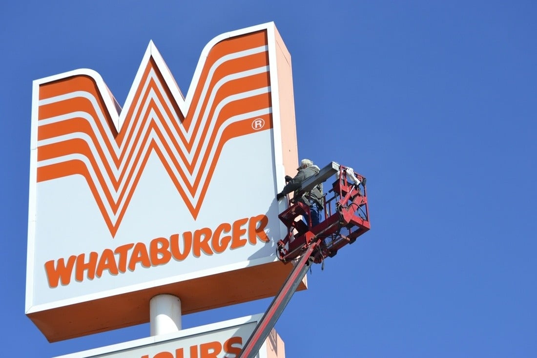 Technician in a bucket lift beside a large orange and white Whataburger pole sign.