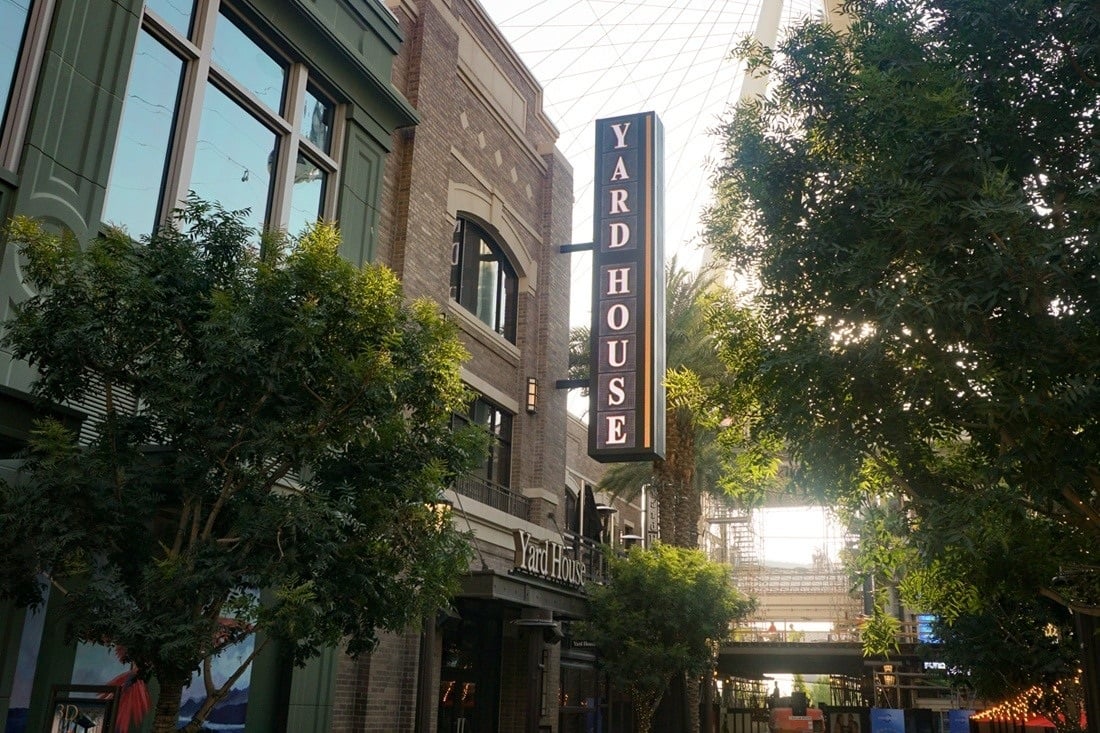Vertical Yard House blade sign mounted on a stone building along a landscaped pedestrian walkway.